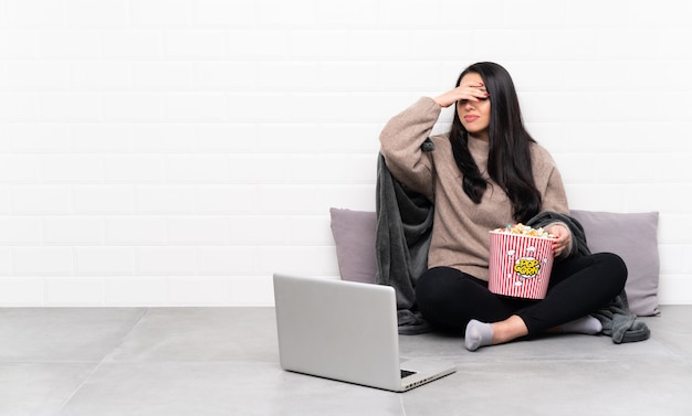 Giovane ragazza colombiana che tiene una ciotola di popcorn e che mostra un film in un computer portatile con espressione stanca e malata
