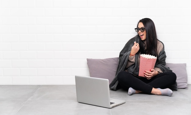 Giovane ragazza colombiana in una stanza con gli occhiali 3d e in possesso di un grande secchio di popcorn mentre guardando di lato