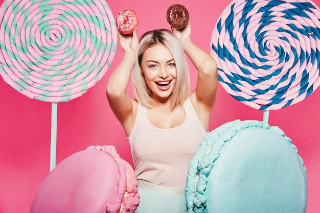 Giovane ragazza con capelli biondi che indossa top e cappello rosa in piedi con enormi lecca-lecca dolci in studio rosa