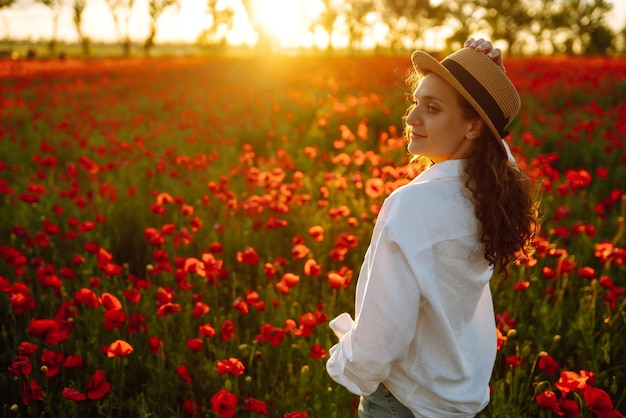 Giovane ragazza con cappello in posa nel campo di papaveri Ritratto di ragazza riccia che cammina nel campo di papaveri al tramonto