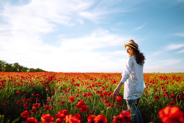 Giovane ragazza con cappello in posa nel campo di papaveri Ritratto di ragazza riccia che cammina nel campo di papaveri al tramonto