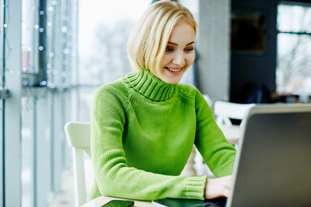 Giovane ragazza con i capelli chiari che indossa un maglione verde seduto nella caffetteria con laptop, ritratto, concetto di freelance, shopping online.