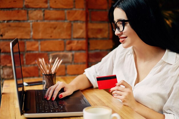 Giovane ragazza con i capelli neri che indossa occhiali seduto in un caffè con laptop, telefono cellulare, carta di credito e tazza di caffè, concetto di freelance, shopping online, indossa una camicia bianca