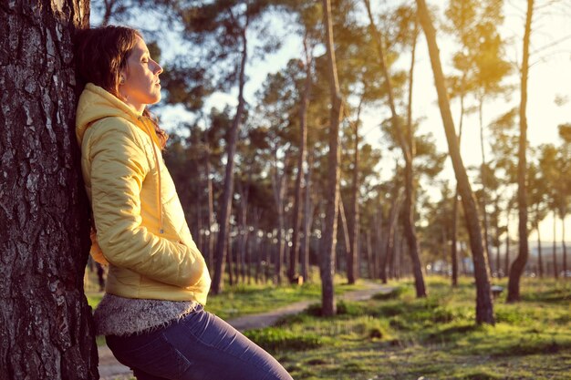 Giovane ragazza con i capelli ricci in giacca gialla e pantaloni blu appoggiata su un pino con gli occhi chiusi godendosi la natura in una foresta di pini al tramonto