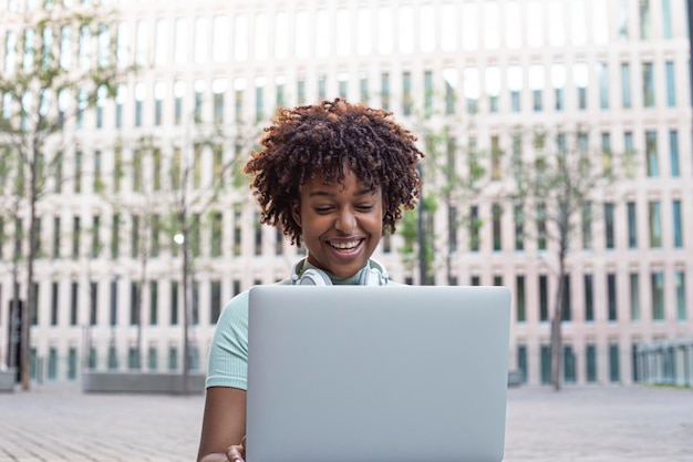 Giovane ragazza con i capelli ricci seduta sulle scale all'aperto e digitando sul suo computer portatile con un sorriso allegro