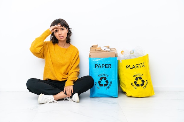 Giovane ragazza con in mano una borsa piena di plastica e carta seduta sul pavimento isolato su sfondo bianco guardando lontano con la mano per guardare qualcosa