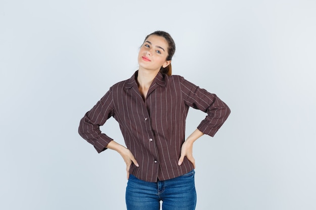 Giovane ragazza con le mani sui fianchi in camicia a righe, jeans e dall'aspetto carino. vista frontale.