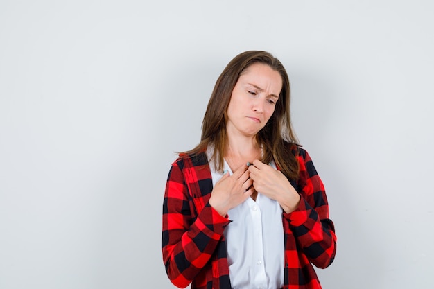 Giovane ragazza con le mani sul petto, labbra inferiori curve in camicia a scacchi, camicetta e sguardo sconvolto, vista frontale.