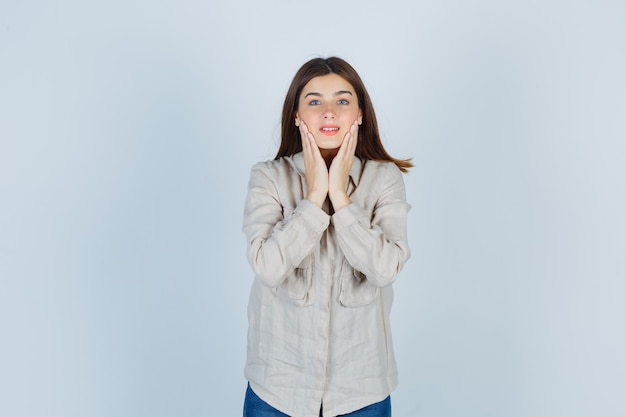 Giovane ragazza con le mani sulle guance, sorridente in camicia beige, jeans e carina, vista frontale.