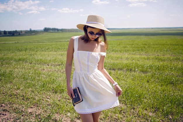 Giovane ragazza con lunghi capelli neri si trova sul sito in abito bianco e vestiti cappello di paglia è sul campo verde con erba e sorrisi, estate.