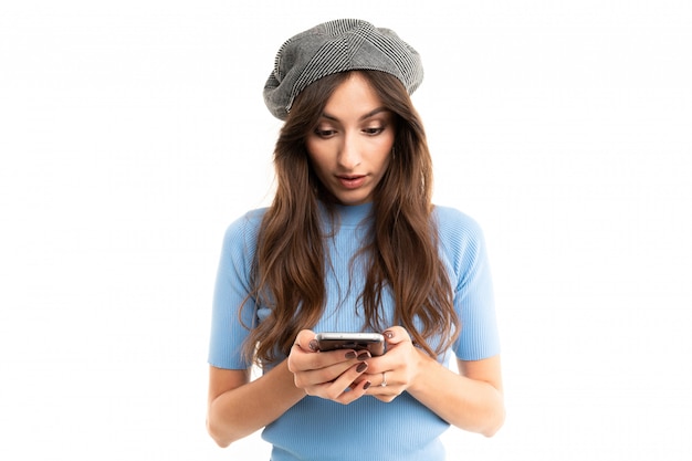 Giovane ragazza con un sorriso delizioso, lunghi capelli castani ondulati, bellissimo trucco, in maglia blu, jeans neri, berretto grigio, con bracciale rosso con telefono in mano