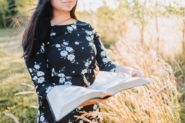 Giovane ragazza cristiana religiosa che tiene e legge la sua bibbia, fuori nel campo al tramonto.