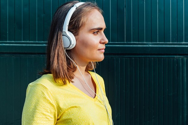 Giovane ragazza dai capelli biondi che indossa una maglietta gialla, ascoltando musica con le cuffie bianche, su sfondo metallico verde. Tecnologia, musica, stile di vita e concetto di relax.