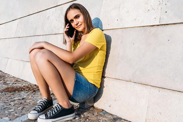 Giovane ragazza dai capelli biondi che indossa una maglietta gialla, parla con il suo smartphone appoggiato a un muro di pietra. Tecnologia, musica, stile di vita e concetto di relax.