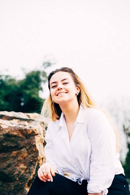giovane ragazza dai capelli lunghi in camicia bianca sorridente seduta su una grande pietra in un parco