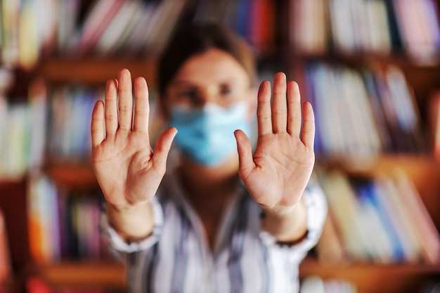 Giovane ragazza del college con la maschera per il viso in piedi in biblioteca e mostrando le mani pulite. Covid pandemia concetto.