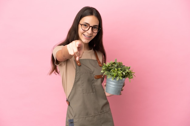 Giovane ragazza del giardiniere che tiene una pianta isolata su fondo rosa che indica la parte anteriore con l'espressione felice