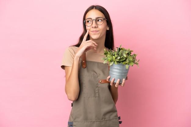 Giovane ragazza del giardiniere che tiene una pianta isolata su fondo rosa che pensa un'idea mentre osserva in su