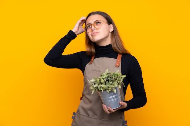 Giovane ragazza del giardiniere che tiene una pianta sopra giallo isolato che ha dubbi e con l'espressione confusa del fronte