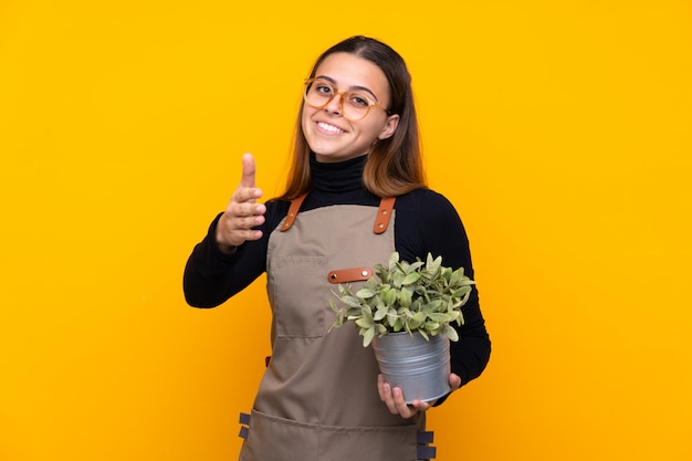 Giovane ragazza del giardiniere che tiene una pianta sopra la stretta di mano gialla isolata dopo il buon affare