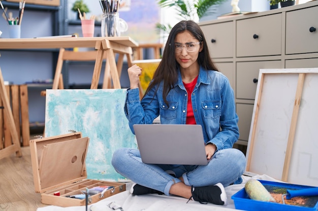 Giovane ragazza dell'adolescente che si siede allo studio d'arte usando il computer portatile infastidito e frustrato gridando con rabbia urlando pazzo con rabbia e mano alzata