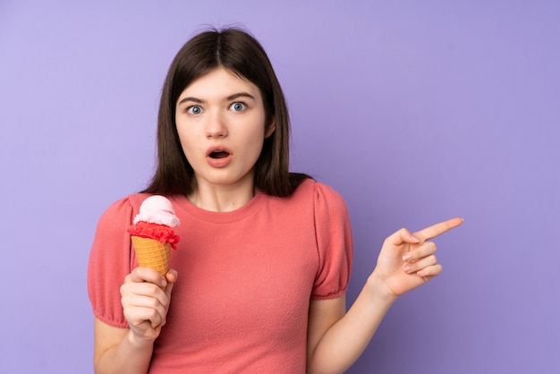 Giovane ragazza dell'adolescente che tiene un gelato della cornetta sopra la parete viola sorpresa e che indica lato