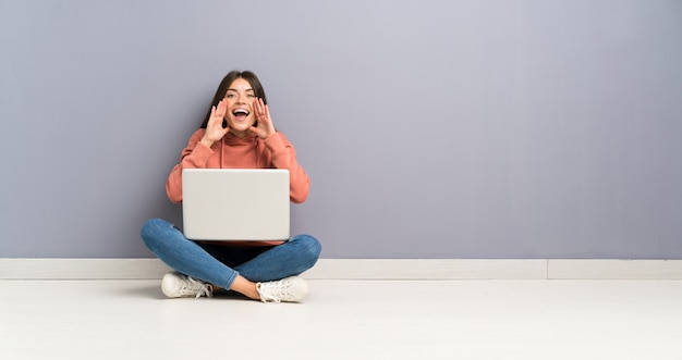 Giovane ragazza dello studente con un computer portatile sul pavimento che grida con la bocca spalancata