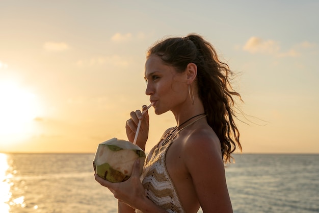 Giovane ragazza di bellezza che beve cocco alla spiaggia tropicale vicino all'acqua di mare all'isola di paradiso al tramonto