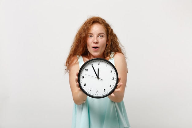 Giovane ragazza di donna rossa stupita eccitata scioccata in vestiti leggeri casuali in posa isolata sul fondo bianco della parete, ritratto in studio. Concetto di stile di vita della gente. Mock up copia spazio. Tenendo l'orologio rotondo.