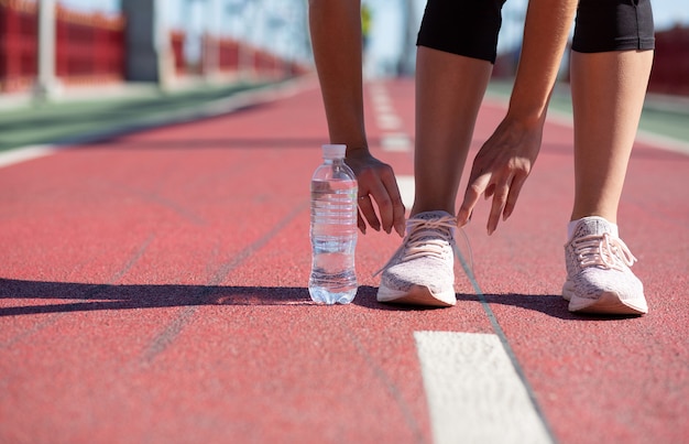 Giovane ragazza di forma fisica che lega i lacci delle scarpe sulla pista da corsa in una giornata di sole. Spazio per il testo