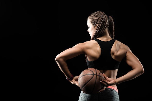 Giovane ragazza di forma fisica con la sfera di pallacanestro dalla parte posteriore su priorità bassa nera