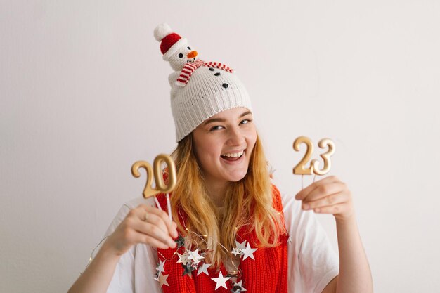Giovane ragazza divertente con un cappello di Capodanno e una sciarpa rossa tiene i numeri 2023 nelle sue mani Concetto di Capodanno e Natale