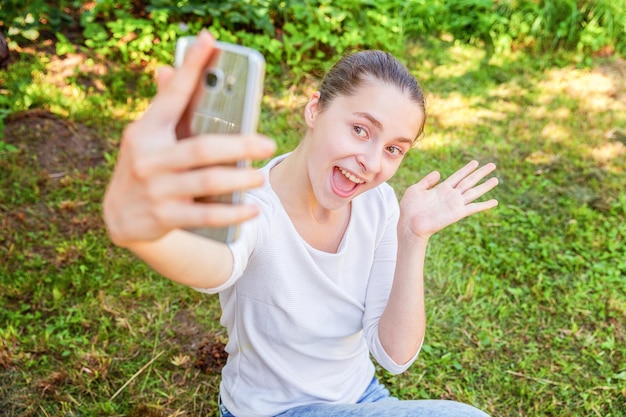 Giovane ragazza divertente prendere selfie dalle mani con il telefono seduto sul parco di erba verde o sullo sfondo del giardino. Ritratto di giovane donna attraente che fa foto selfie su smartphone nel giorno d'estate