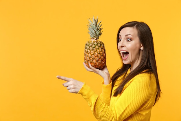 Giovane ragazza eccitata che tiene la bocca aperta puntando il dito indice da parte tenendo frutta fresca di ananas isolata su sfondo giallo arancione. Lo stile di vita vivido delle persone si rilassa il concetto di vacanza. Mock up copia spazio