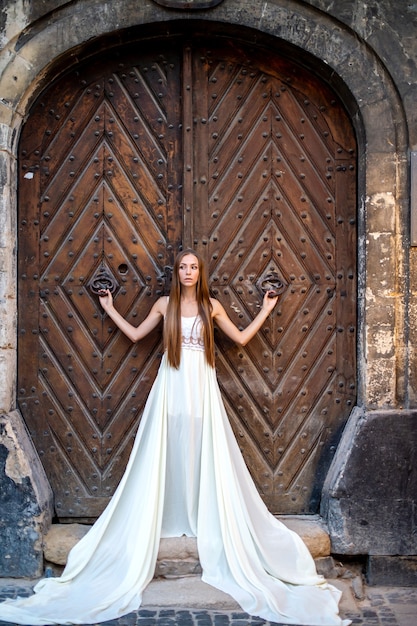 Giovane ragazza elegante romantica in vestito bianco lungo che posa sopra la porta antica