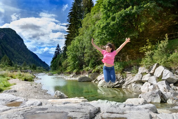Giovane ragazza energica che celebra la sua libertà saltando in aria con le braccia tese sulle rocce lungo un fiume in una valle di montagna
