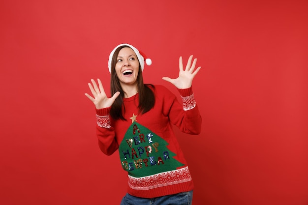 Giovane ragazza felice allegra di Santa in maglione lavorato a maglia Cappello di Natale che cerca le dita di diffusione delle mani isolate su fondo rosso. Felice Anno Nuovo 2019 celebrazione festa concetto. Mock up copia spazio.