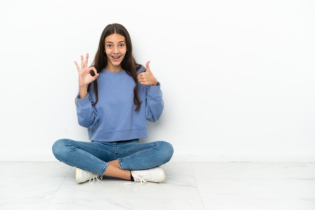 Giovane ragazza francese che si siede sul pavimento che mostra segno giusto e pollice sul gesto