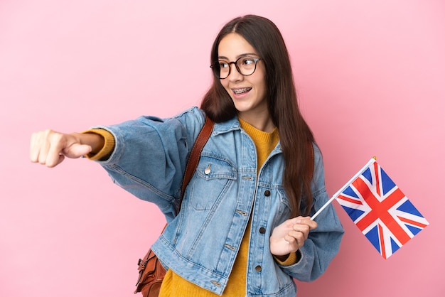 Giovane ragazza francese che tiene una bandiera del Regno Unito isolata su fondo rosa che dà un pollice in alto gesto