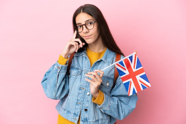 Giovane ragazza francese che tiene una bandiera del Regno Unito isolata su sfondo rosa pensando un'idea