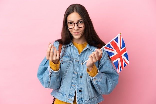 Giovane ragazza francese con in mano una bandiera del Regno Unito isolata su sfondo rosa che fa gesti di denaro
