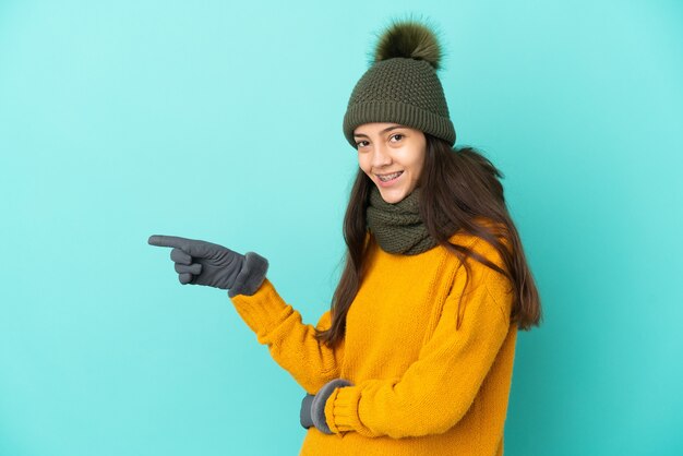 Giovane ragazza francese isolata su sfondo blu con cappello invernale che punta il dito a lato