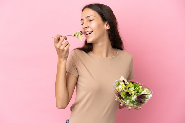 Giovane ragazza francese isolata su sfondo rosa, tenendo una ciotola di insalata e guardandola con felice espressione