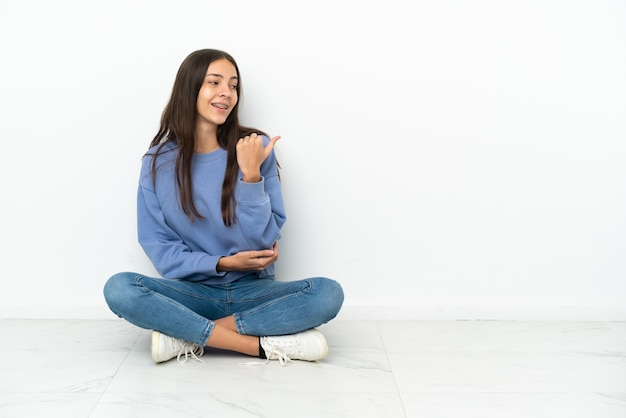 Giovane ragazza francese seduta sul pavimento che punta di lato per presentare un prodotto