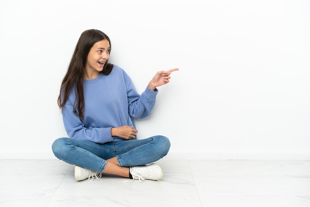 Giovane ragazza francese seduta sul pavimento che punta il dito di lato e presenta un prodotto