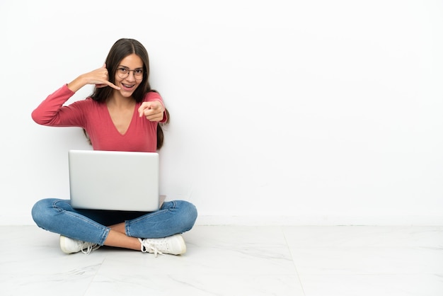 Giovane ragazza francese seduta sul pavimento con il suo computer portatile che fa il gesto del telefono e indica la parte anteriore