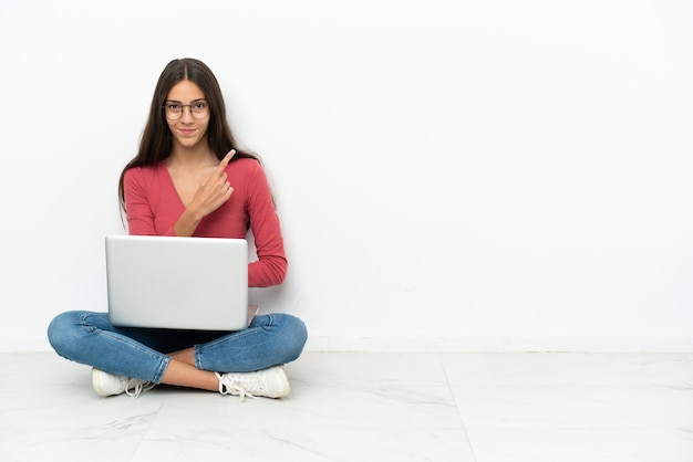 Giovane ragazza francese seduta sul pavimento con il suo laptop rivolto verso il lato per presentare un prodotto