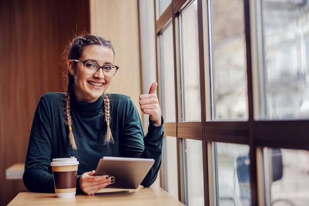 Giovane ragazza geeky sorridente che si siede nella caffetteria, godendo del tempo libero, tenendo il tablet e mostrando i pollici in su. Nuovo concetto di tecnologie.