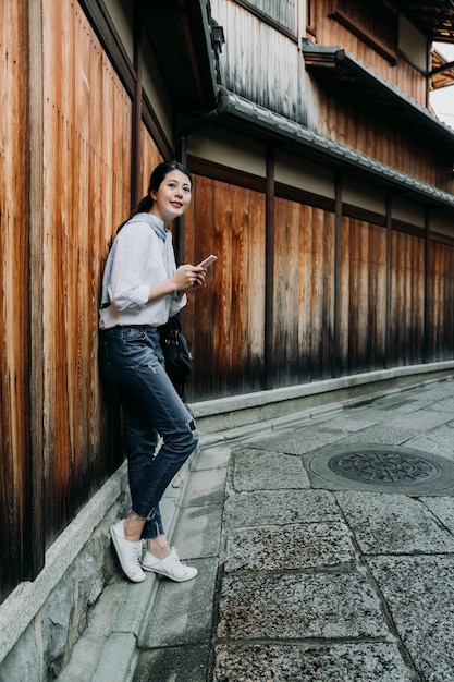 giovane ragazza giapponese che tiene il telefono cellulare facendo affidamento sulla parete di legno in attesa di un amico. donna asiatica sorridente che utilizza un messaggio di sms sul cellulare in chat online. bella signora percorso rilassante modo ishibe vicolo