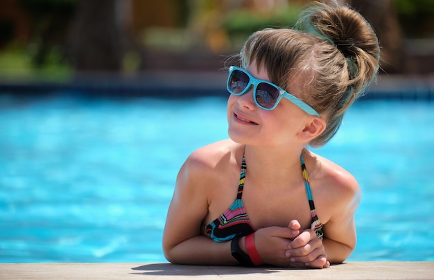 Giovane ragazza gioiosa del bambino che riposa sul lato della piscina con acqua azzurra chiara nella soleggiata giornata estiva Concetto di vacanze tropicali